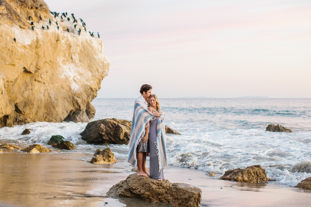 The couple wrapped in a blanket on a rock. 